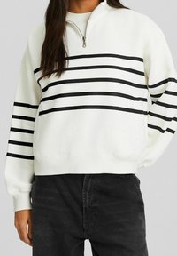 Felpa bianca con collo alto e zip a un quarto, caratterizzata da ampie strisce orizzontali nere e polsini a coste, abbinata a jeans scuri.