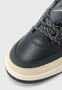 Σκού�ρο δερμάτινο αθλητικό παπούτσι με υφή στην επάνω επιφάνεια, μπεζ σόλα από σουέντ, και αντίθετα πράσινα ακροφύσια με κορδόνια με σχέδια και λεία μύτη.