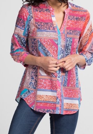 Femme portant une chemise à motifs colorés à manches longues avec des boutons et un jean bleu, les mains jointes à la taille.