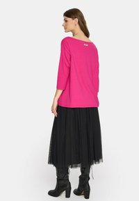 Fuksjowa bluzka z długim rękawem i szerokim dekoltem, zestawiona z czarną tiulową spódnicą i czarnymi wysokimi kozakami na obcasie. Metka jest widoczna z tyłu.