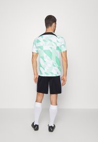 Herrernas sportkläder med en kortärmad skjorta i mintgrön med geometriskt mönster, svarta shorts, vita knälånga strumpor och svarta fotbollsskor.