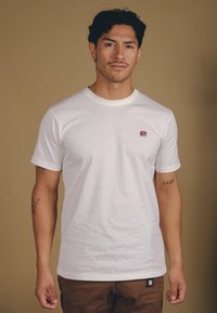 Weißes Baumwoll-T-Shirt mit Rundhalsausschnitt, kurzen Ärmeln und einem kleinen roten Elefanten-Logo auf der linken Brust. Das Modell trägt beigefarbene Hosen.