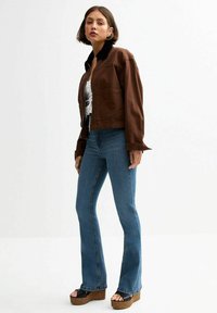 Brun kort jacka med en svart krage, kombinerad med utsvängda blå jeans och bruna plattformssandaler. Modellen står mot en enfärgad bakgrund.