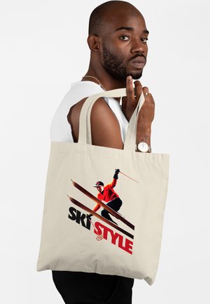 Man in wit mouwloos shirt met een beige draagtas met een kleurrijke skiër en de tekst "SKI STYLE" tegen een effen achtergrond.