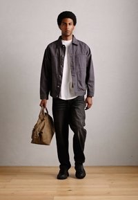 Mann, der drinnen geht, trägt eine graue Jacke, ein weißes Hemd, schwarze Hosen, schwarze Schuhe und hält in seiner linken Hand eine braune Ledertasche.