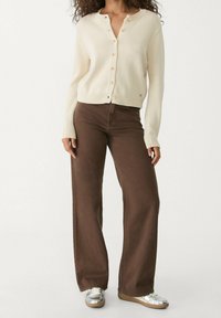 Kremowy, dziany kardigan z okrągłym dekoltem i zapięciem na guziki, zestawiony z brązowymi spodniami o szerokiej nogawce i srebrnymi sneakersami. Teksturowany materiał i luźny krój.