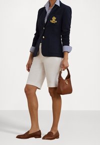 Person klädd i en marinblå blazer med guld-emblem, randig skjorta, vita knälånga shorts, bruna loafers och som håller en liten brun handväska.