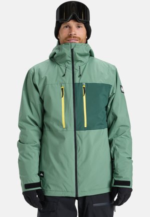 Groene jas met een donkergroen borstpaneel, gele ritsen, ritszakken, verstelbare capuchon en zwarte handschoenen. Gladde nylon textuur.