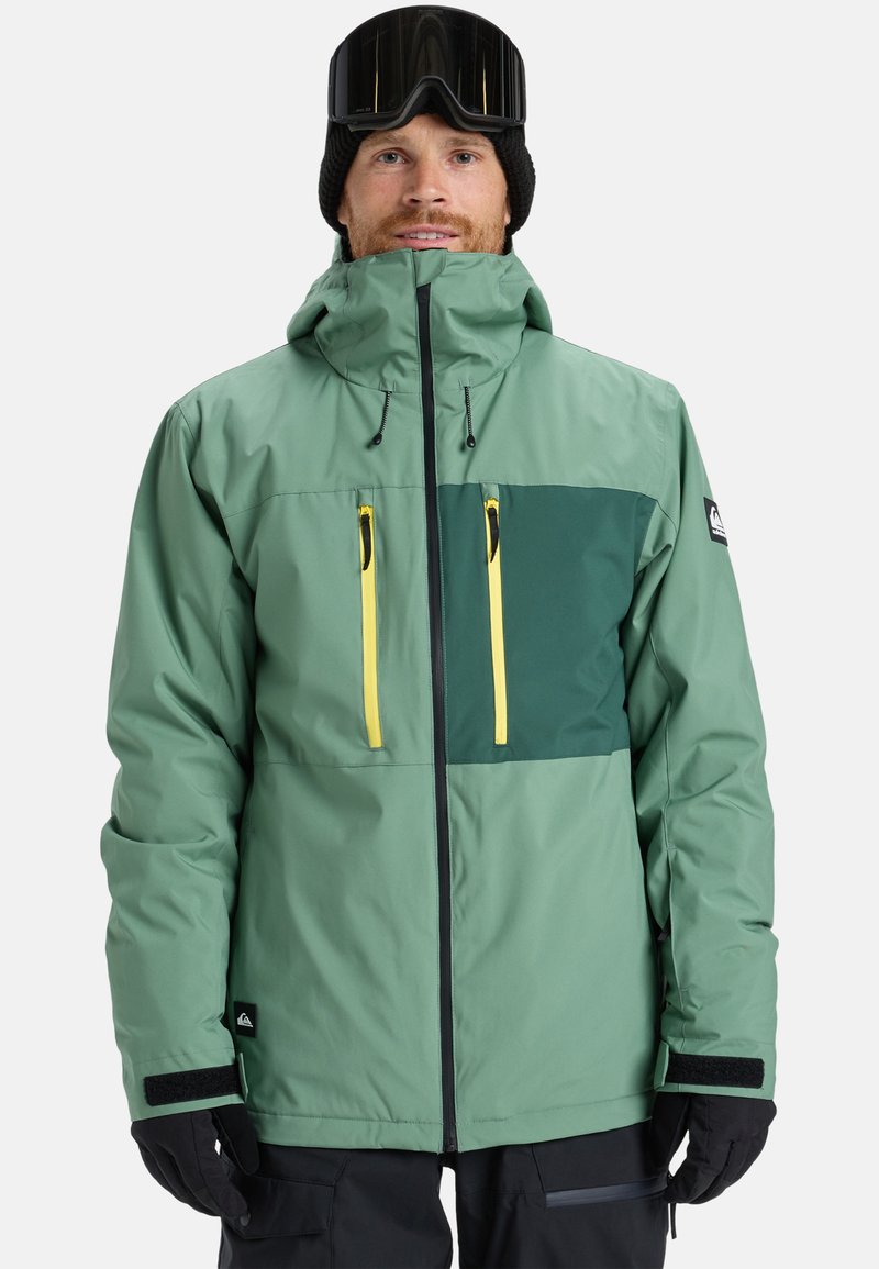 Grøn jakke med en mørkegrøn brystpanel, gule lynlåse, lynlåslommer, justerbar hætte og sorte handsker. Glat nylon tekstur.