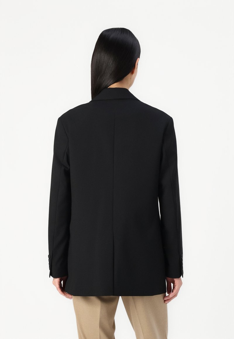 Schwarzer taillierter Blazer mit Reverskragen, einzelnen Rückenschlitz und langen Ärmeln, kombiniert mit beigen Hosen. Glatte Textur, formelles Design.