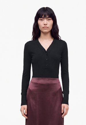 Donna con lunghi capelli neri che indossa una maglia nera a maniche lunghe con scollo a V e bottoni e una gonna lucida viola scuro, in piedi davanti a uno sfondo bianco.