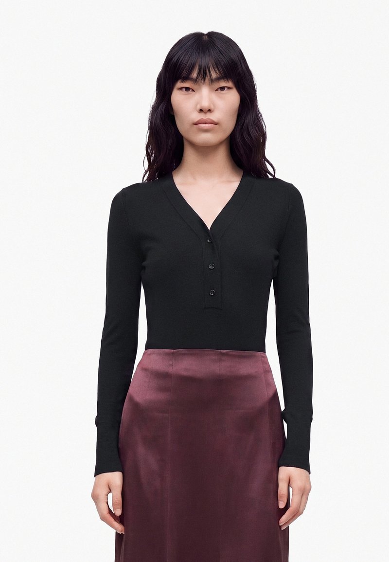 Femme aux longs cheveux noirs portant un haut noir à manches longues en col V avec des boutons et une jupe brillante violet foncé, debout devant un fond blanc.