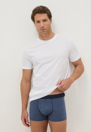 Homme portant un t-shirt blanc à manches courtes le soulève légèrement pour montrer un boxer bleu à petits pois blancs sur un fond clair uni.