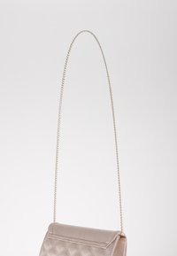 Sac à main matelassé beige avec une bandoulière en chaîne dorée, doté d'un rabat et d'une texture douce. Design minimaliste et forme compacte.