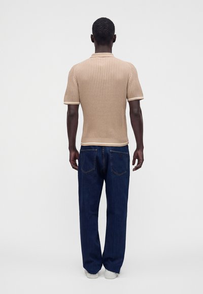 Homme debout de dos portant un t-shirt côtelé beige à manches courtes, un jean bleu foncé et des baskets blanches sur un fond clair uni.