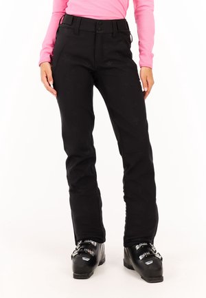 Kobieta ubrana w czarne spodnie narciarskie i czarne buty narciarskie, stojąca na białym tle.