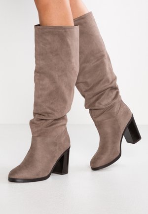 Bottes mi-mollet en daim taupe avec tiges larges et talons blocs noirs massifs portées sur jambes croisées sur fond blanc uni.