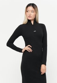 Črna dolga obleka z dolgimi rokavi in polovičnim zadrgo na ovratniku, izdelana iz mehkega rebrastega materiala. Značilnost je majhen logotip na prsih in prilegajoč silhuet.