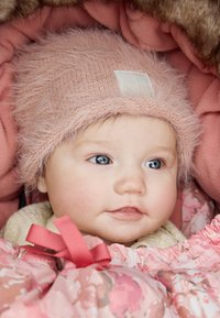 Ljusrosa fluffig hatt med en fyrkantig etikett, bebis i en blommönstrad rosa filt med volangkanter, mjuka texturer och rosetter. Ljus hy och blå ögon.