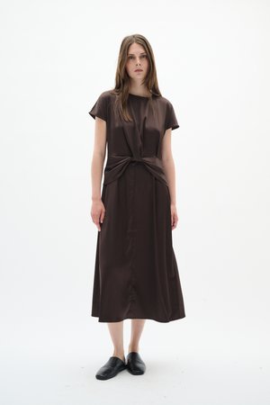 Naine sirgete pruunide juustega, seljas pruuni satiinist keskmise pikkusega kleit, millel on talje juures lipsuga detail, ning mustad sussid, taustaks valge sein.