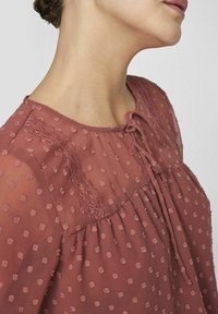Rosa, genomskinlig blus med prickmönster, spetsdetaljer vid halsringningen och knutstängning. Har en avslappnad passform och mjuk textur.