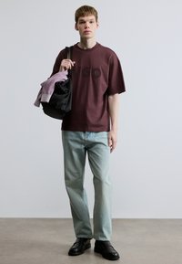 Oversized Bordeaux t-shirt med præget logo, lyseblå jeans, holder en sort taske og en foldet lilla sweater, kombineret med sorte sko.