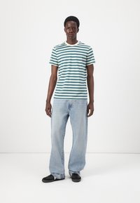 Striped t-shirt in teal and white, short sleeves, crew neck. Worn with light blue baggy jeans and black sneakers. Minimalist background.