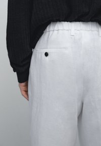Weiße Leinenhosen mit entspanntem Schnitt, ausgestattet mit einem elastischen Bund und einer einzelnen aufgesetzten Tasche mit einem schwarzen Knopfdetails.