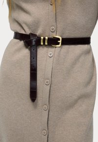 Ceinture en cuir brun foncé avec boucle dorée attachée sur une robe en maille beige boutonnée.