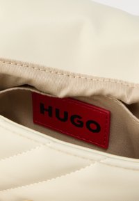 Beež sünteetilise käekoti sisemus, millel on tikitud tekstuur ja silmapaistev punane "HUGO" silt. Sile materjal peene läikega.