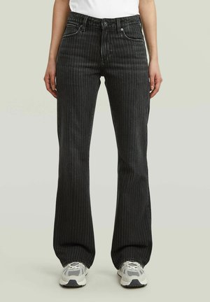 Jeans a zampa con righe nere indossati con sneakers bianche e una maglietta bianca, mostrati dalla vita ai piedi su uno sfondo neutro.