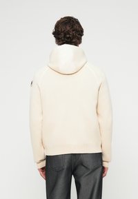 Άτομο με σγουρά μαλλιά που φοράει κρεμ πουλόβερ με κουκούλα και σκούρο γκρι τζιν, γυρισμένο πλάτη μπροστά σε λευκό απλό φόντο.