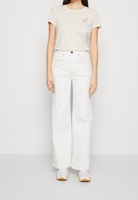 Camiseta de algodón beige con bordado de hoja, combinada con jeans blancos de pierna ancha de talle alto y zapatillas grises. Conjunto sencillo y casual.