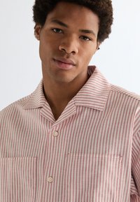 Mladý muž s kučeravými vlasmi, ktorý nosí ružovú a bielu košeľu s vertikálnymi pruhmi na gombíky, stojaci pred jednoduchým pozadím.