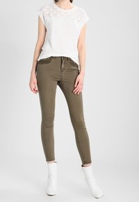 Bílý tričko s texturovanou krajkovou částí, kombinované s olivově zelenými skinny džíny a bílými kotníkovými botami. Jednoduchý, přiléhavý design.