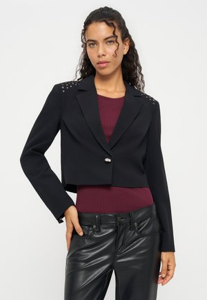 Jeune femme aux cheveux bouclés portant un blazer court noir à épaules cloutées, un haut bordeaux et un pantalon en cuir noir.