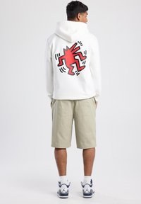 Sudadera blanca con un gráfico rojo y negro en la parte posterior, combinada con pantalones cortos de caqui claros y zapatillas blancas con acentos azules.