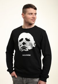 Felpa nera con una grande grafica bianca di una maschera sul davanti e il testo "JOHN CARPENTER'S HALLOWEEN" sotto il design.