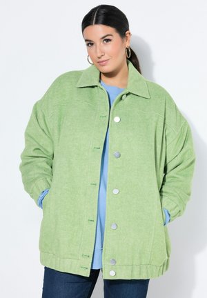Donna con capelli scuri raccolti in una coda di cavallo, indossa orecchini a cerchio dorati, una giacca oversize verde chiaro, una camicia blu e jeans scuri, con le mani nelle tasche.