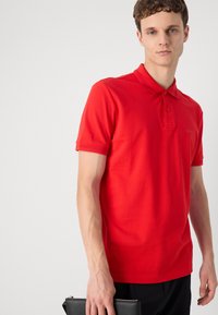 Joven vestido con una camisa polo roja y pantalones negros, sosteniendo un bolso de mano negro frente a un fondo blanco liso.