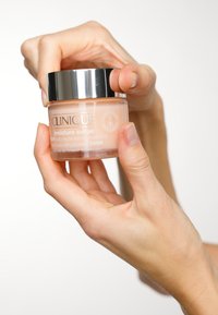 Clinique Moisture Surge purkki, läpinäkyvä vaalean persikan värinen kerma, hopeinen kansi, pyöreä muoto; käsi pitämässä sitä näyttää kiiltävän pinnan ja etikettitiedot.