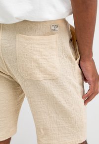 Beige strukturierte Shorts mit einer sichtbaren hinteren Tasche und einem kleinen Markenschild. Der Stoff hat ein gewebtes, gemustertes Aussehen.