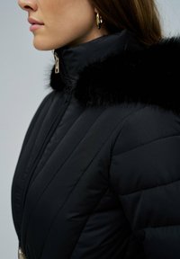 Chaqueta acolchada negra con cuello alto adornado con ribete de piel sintética negra, diseño acolchado vertical y cierre con cremallera de tono dorado.