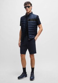 Námornícky puffer vest s horizontálnym prešívaním a detailom loga, v kombinácii s námorníckymi šortkami a tmavými teniskami. Hladká látka s priliehavým strihom.