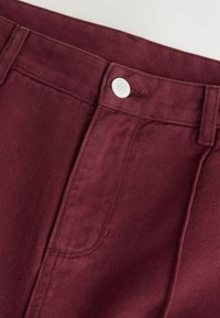 Bordeaux bukser med metalknaplukning, subtil ribbet tekstur og sydetalje langs taljen og sidelommerne.