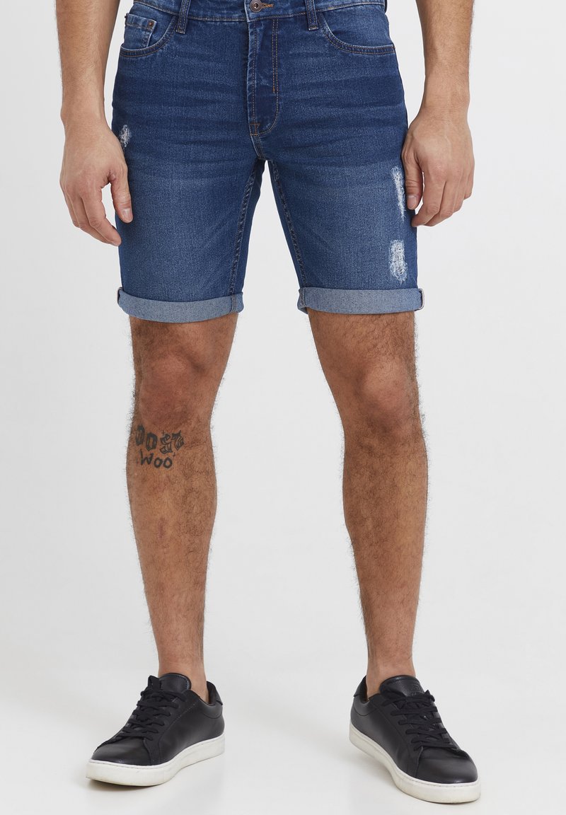Denimshorts i mørk blå med slitte detaljer og oppbrettede kanter. Kombinert med svarte joggesko med hvite såler.