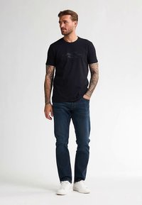 Zwart katoenen T-shirt met een ronde hals en een frontafbeelding, gecombineerd met donkerblauwe jeans en witte sneakers. Model staand tegen een lichte achtergrond.