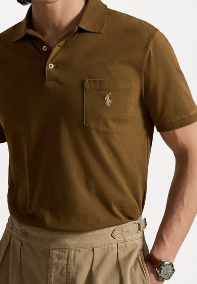 Brown cotton polo shirt with a three-button placket, embroidered logo above the left pocket, paired with pleated beige trousers and a watch.