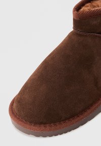Nærbilde av brune semskede vinterstøvler med tekstilkant og gummisåle mot en lys, ensfarget bakgrunn.