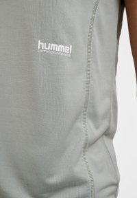 Lys grå ærmeløs træningsskjorte med en tekstureret stof, der har et fremtrædende hvidt logo "hummel PERFORMANCE" på forsiden.